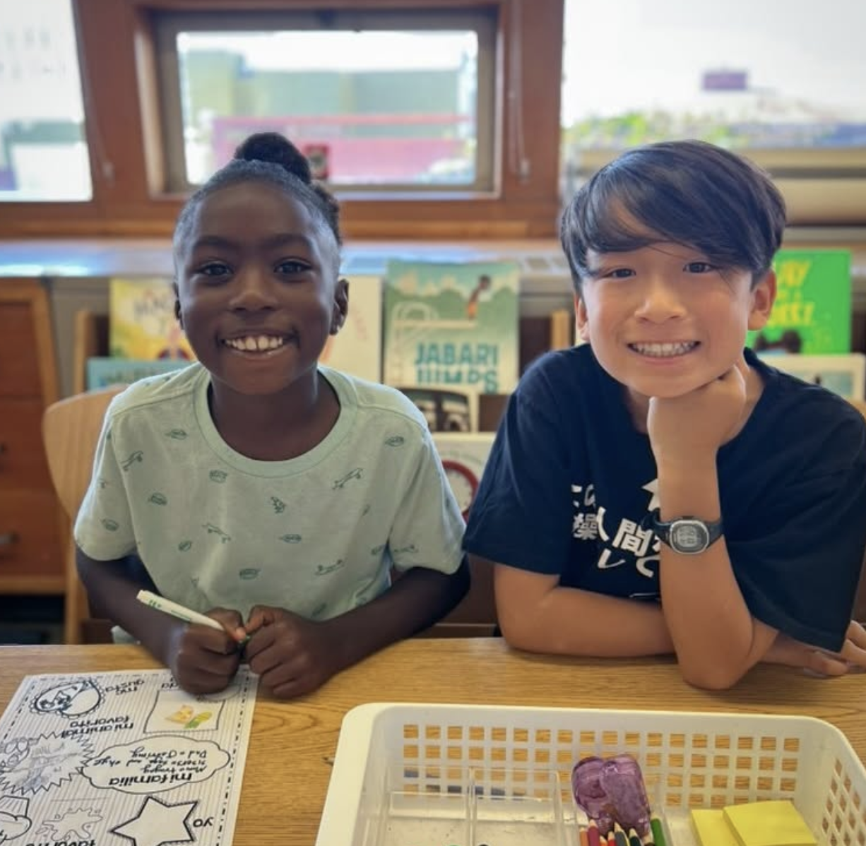 Two people sit together at a desk, smiling at the camera. One holds a marker over a coloring sheet, while the other rests their chin on their hand. Books and supplies are visible in the background.