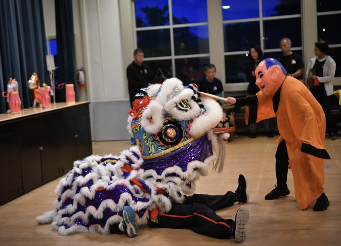 A person in a vibrant lion costume performs a traditional lion dance indoors as another in an orange robe and mask interacts playfully. Several people watch in the background.