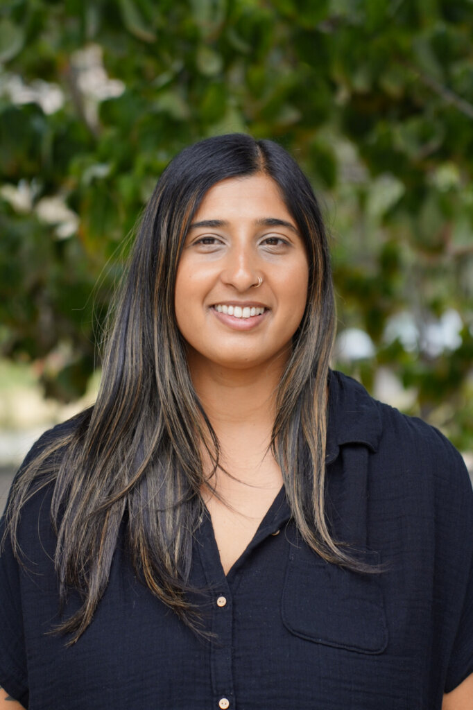 A person with long, straight dark hair and a nose ring smiles at the camera, wearing a black shirt in front of green foliage.