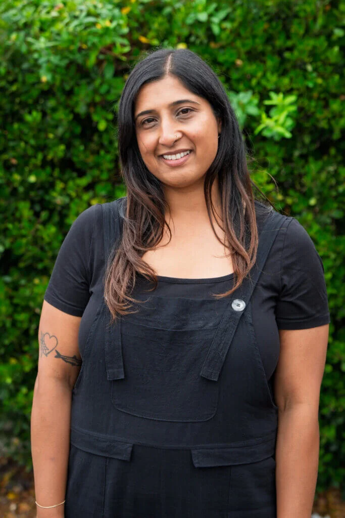 A person with long dark hair, wearing a black shirt and overalls, stands outdoors by green bushes, smiling softly. A heart tattoo is on their arm.
