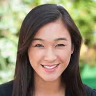 A person with long straight dark hair smiles at the camera, wearing a black top outdoors with blurred green foliage in the background.