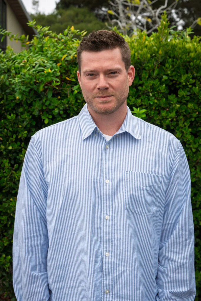 A person stands outdoors in front of a green leafy hedge, wearing a light blue and white striped button-up shirt, looking at the camera.