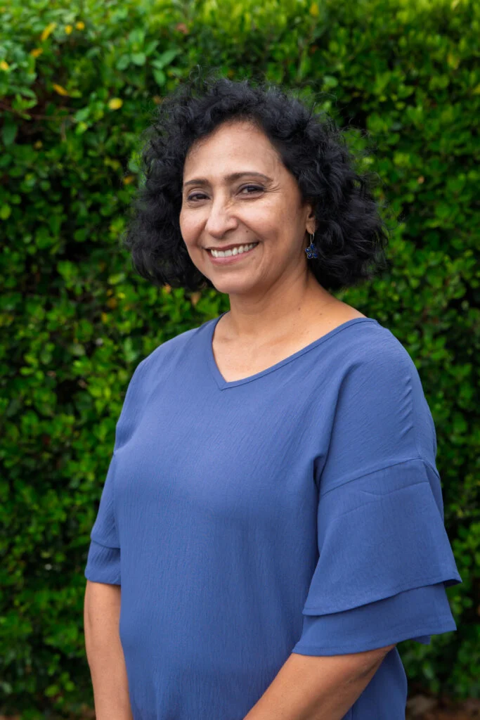 A person with curly black hair, wearing a blue blouse, smiles while standing outdoors in front of green bushes.