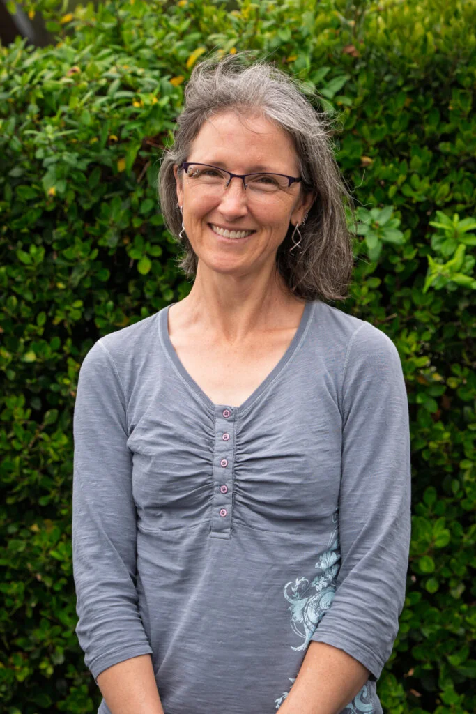 A person with gray hair and glasses, wearing a gray shirt, stands outdoors smiling in front of green bushes.