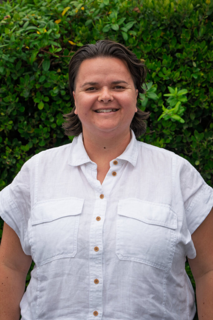 A person with short brown hair in a white button-up shirt stands smiling in front of green leafy bushes.