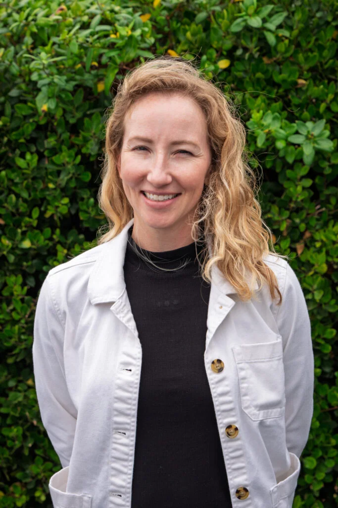 A person with wavy blonde hair, wearing a white jacket over a black top, stands before a green leafy background, smiling.