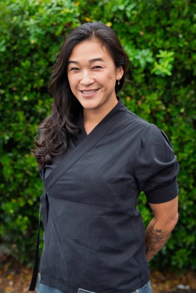 A person with long dark hair, wearing a black top, stands outdoors by green bushes, smiling with hands behind their back.