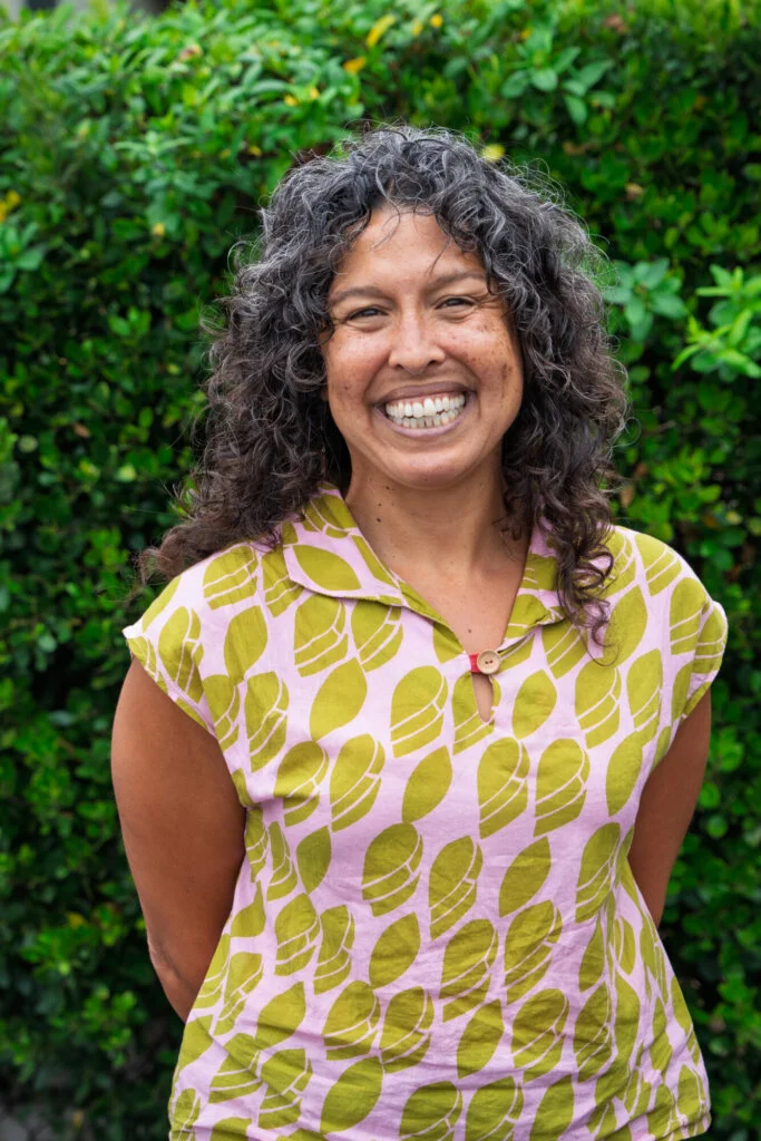 A person with curly hair smiles at the camera, standing in front of green bushes, wearing a pink and green patterned shirt.