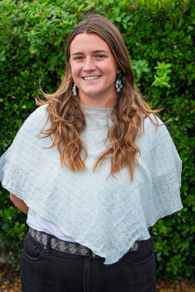A person with long brown hair, wearing a light blue shawl, white top, black jeans, and blue earrings, smiles by a green hedge.