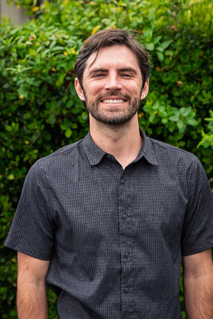 A person with short dark hair and a beard, wearing a short-sleeved dark gray shirt, stands smiling in front of green leafy bushes.