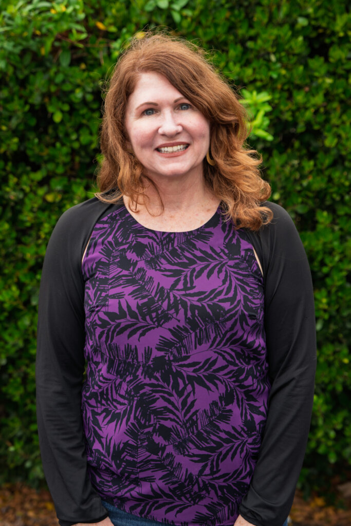 A person with wavy auburn hair stands smiling in front of green bushes, wearing a purple and black long-sleeve shirt with a leaf pattern.