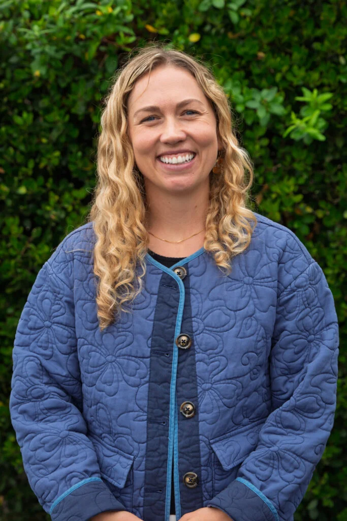 A person with long, curly blond hair smiles while wearing a blue quilted jacket, standing outdoors before a leafy green hedge.