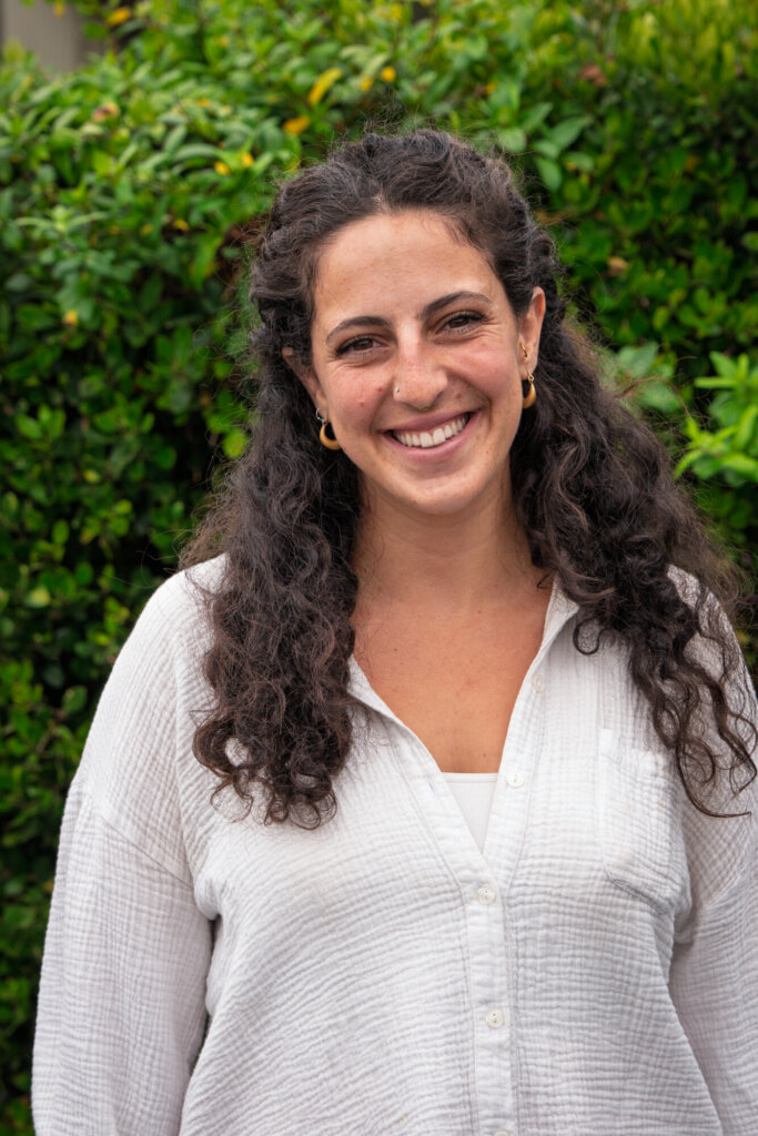 A person with long curly hair, wearing a white shirt, stands outdoors in front of green bushes, smiling at the camera.