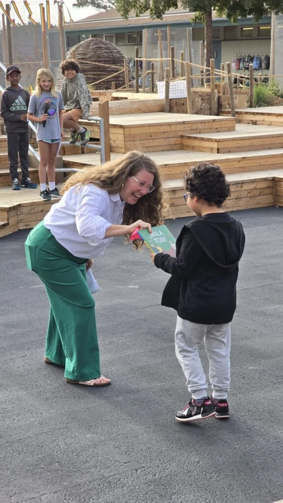 A person with long hair hands a book to another person outdoors, both smiling. Others stand and sit on wooden steps in a schoolyard setting.