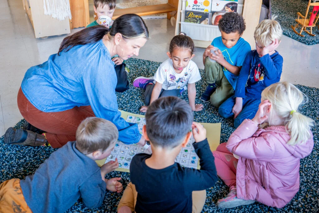 An adult sits on the floor with six children, engaging them in an educational board game in a bright classroom.