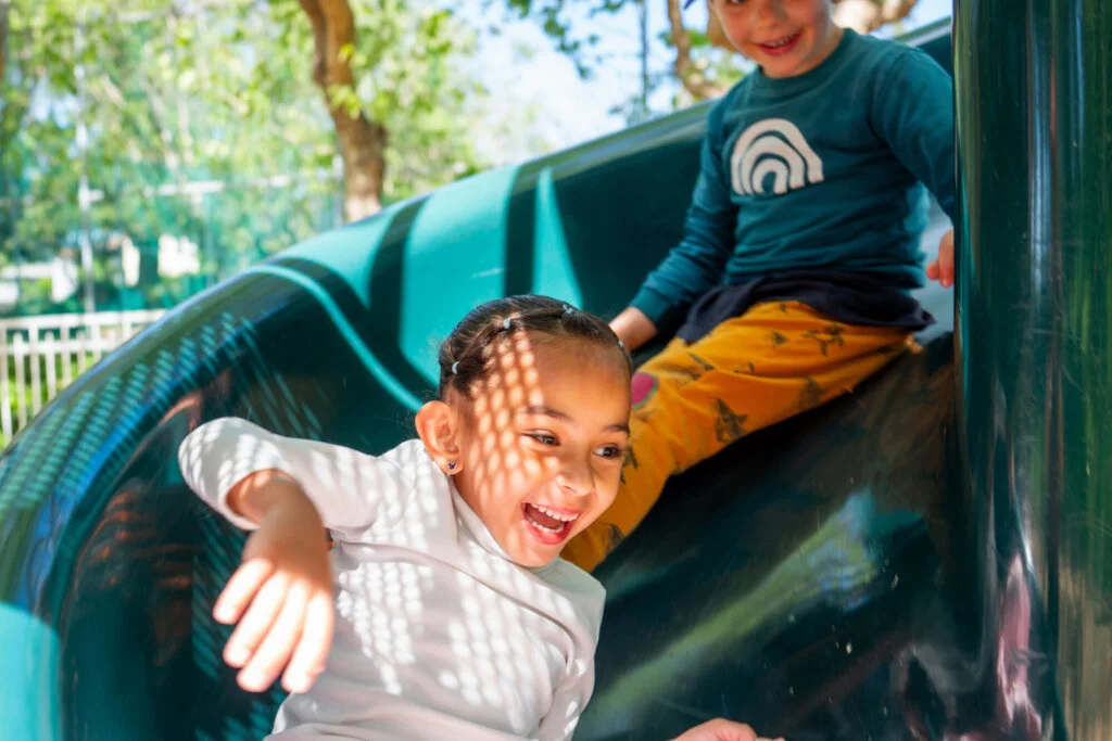 Two smiling people play on a green slide in a sunlit park. Dappled light falls on their faces. Trees and a fence are in the background.