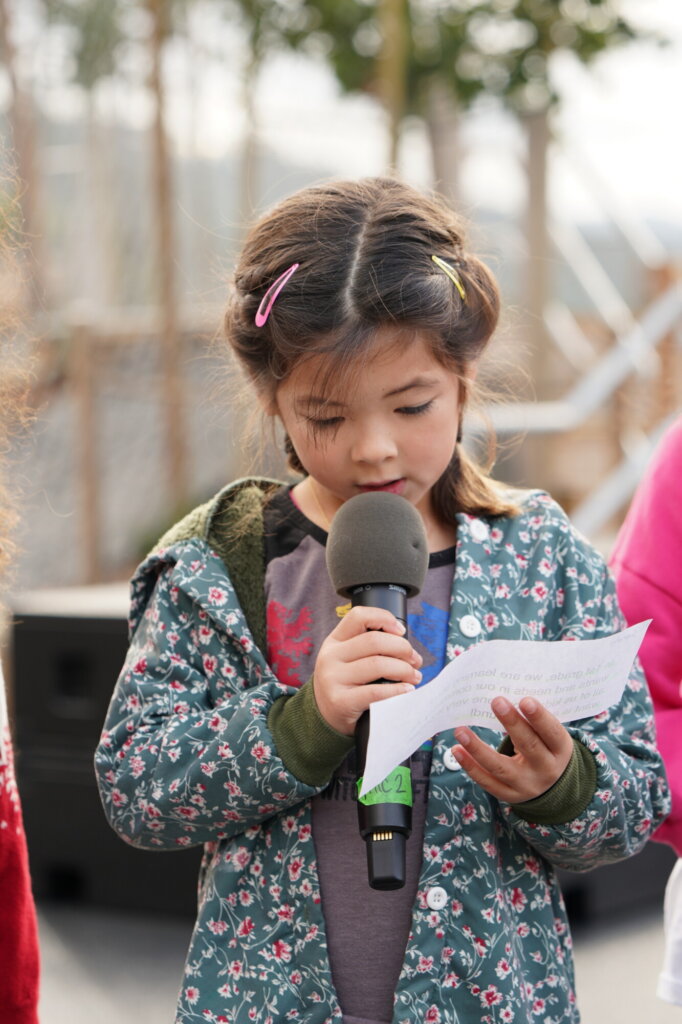 Person in a green patterned jacket holds a microphone and reads from a piece of paper outdoors. Two others are partially visible beside them.