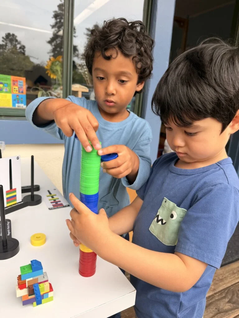 Two people stack colorful plastic disks on a table, building a tall tower. Toys and educational materials are in the background near a window.