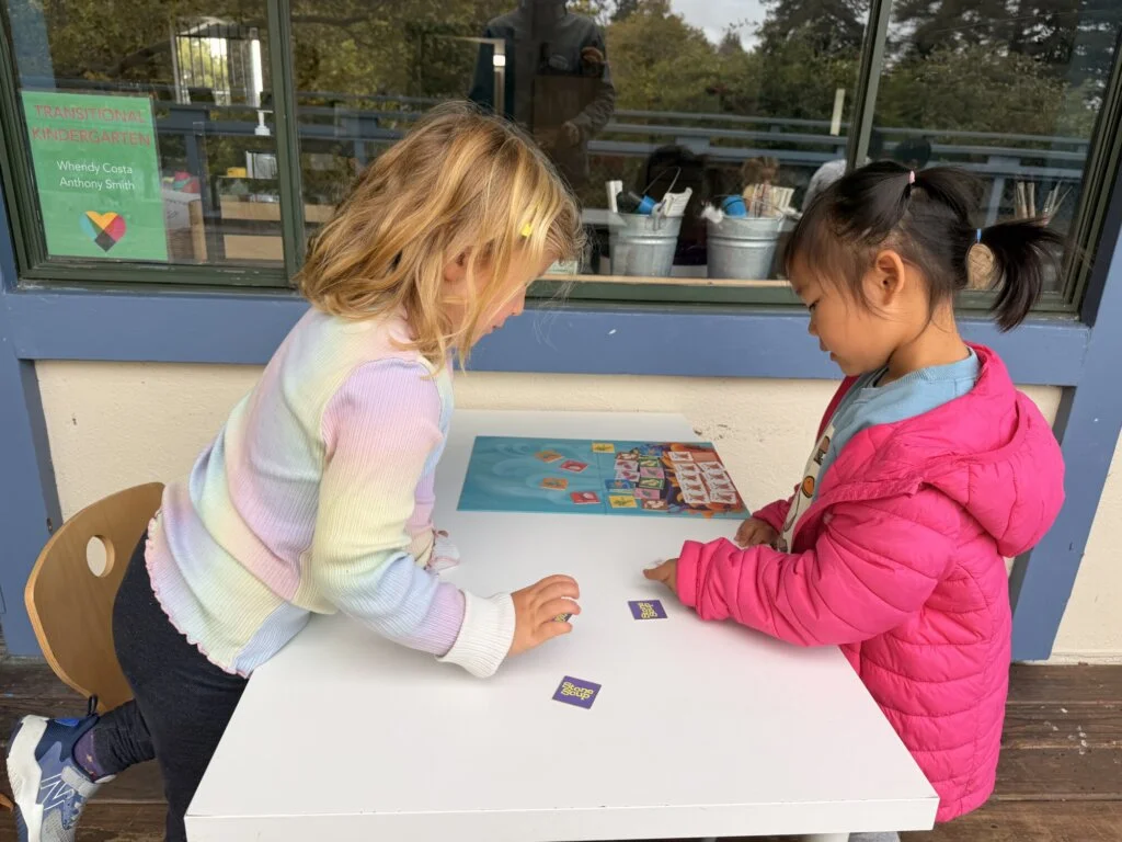 Two people sit at a small white table, playing a matching card game. One wears a pink jacket, the other a pastel sweater. Craft supplies and a sign are visible in the background.