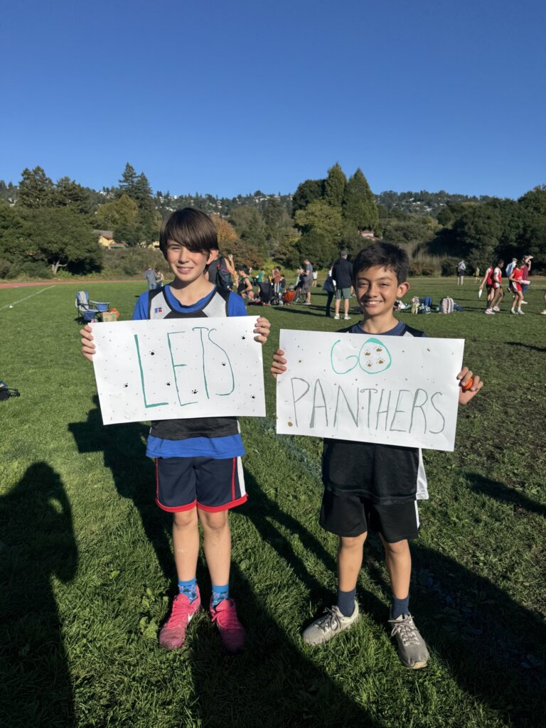 Two people stand outside on grass, smiling and holding signs that say LETS and GO PANTHERS with a soccer ball drawn on one. Other people and trees appear in the background under a clear blue sky.