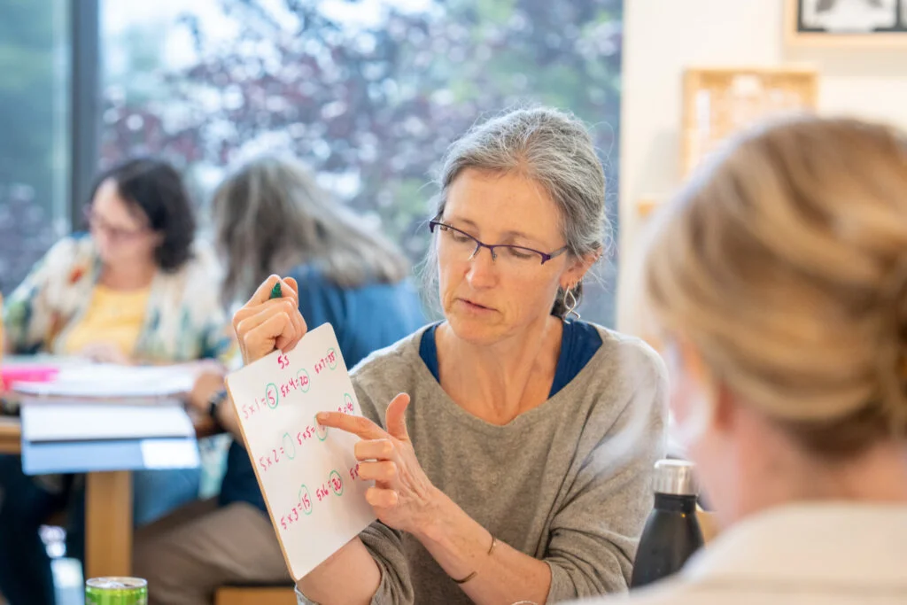 A person wearing glasses points to math equations on a whiteboard while explaining to another, with two more people in the bright classroom.