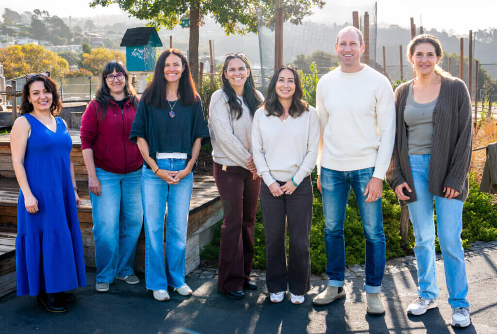 Seven people stand outdoors, smiling at the camera. They wear casual clothing on pavement with greenery, trees, and a wooden bench behind.