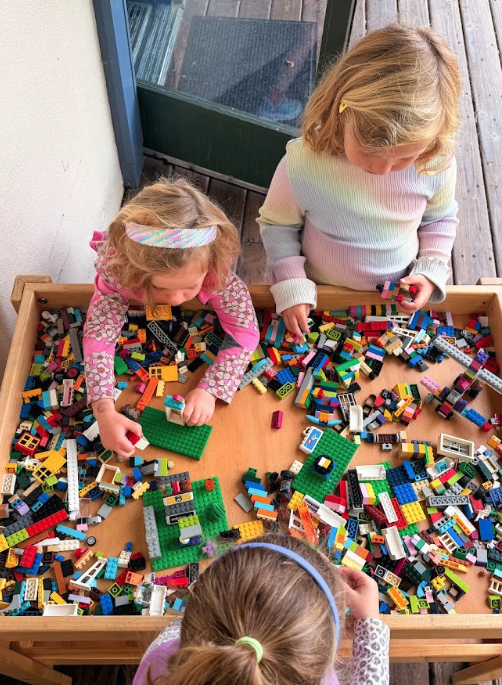 Three people play with colorful LEGO bricks on a wooden table, focused on building. The scene is indoors near a window or door.