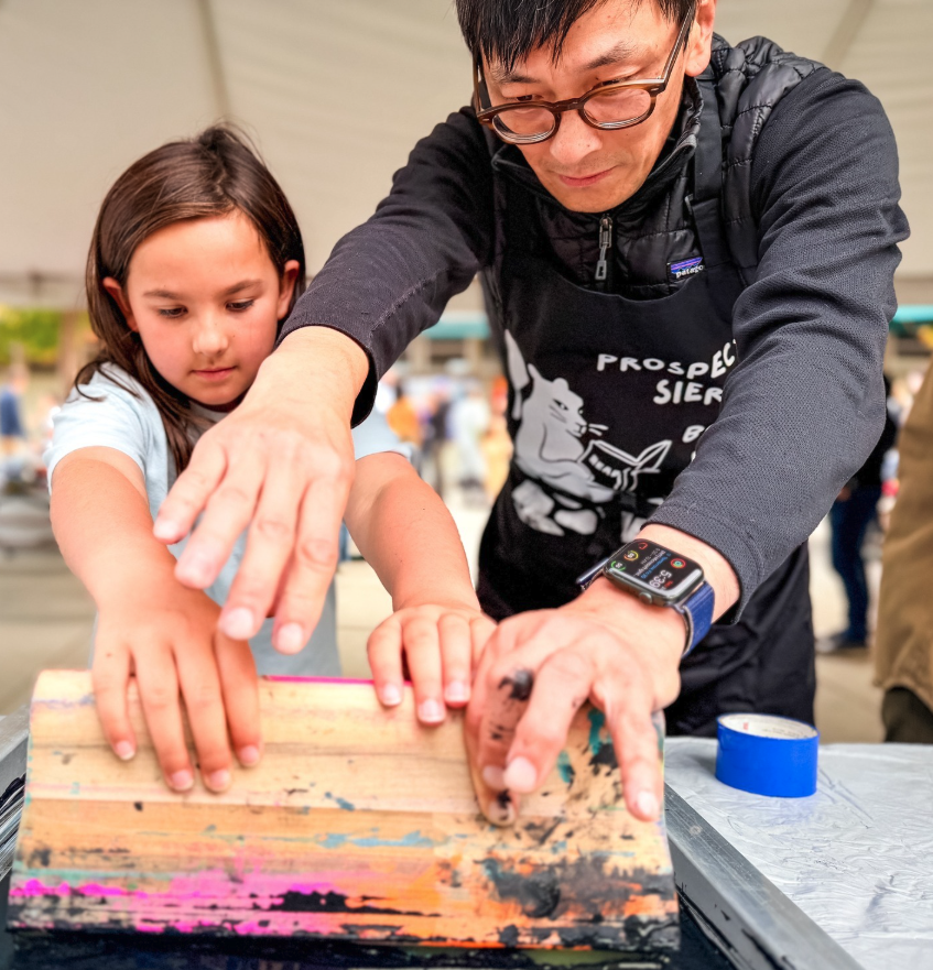 Two people wearing aprons work together to press a screen printing squeegee onto a surface.