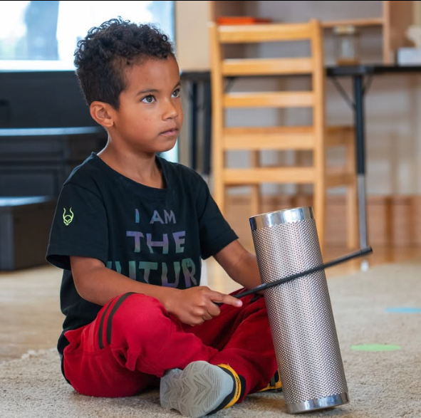 A person sitting on a floor playing a metal guiro percussion instrument.