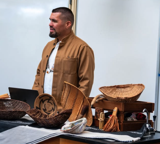 A person stands behind a table displaying woven baskets and an abalone shell.